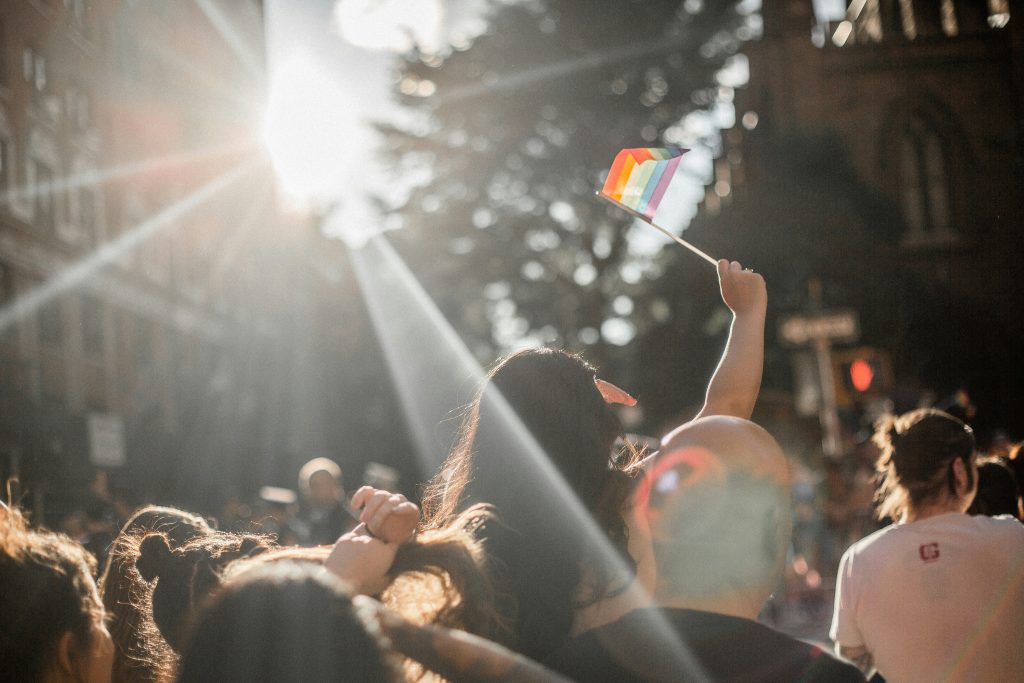 A photo taken from within a crowd of people in a street scene with shards of sunlight shining down and a woman holding a rainbow flag